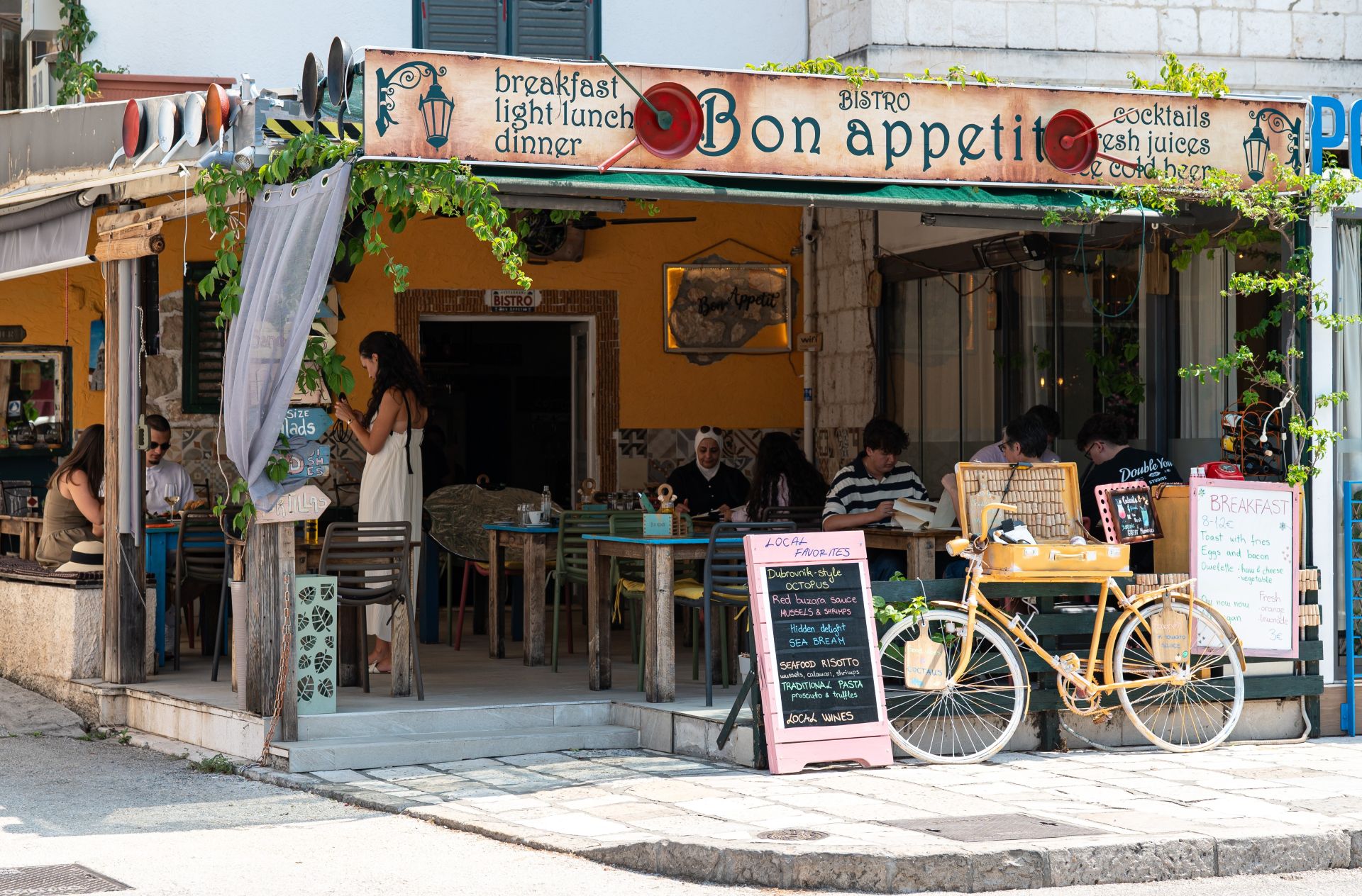 The cheerful yellow front of Bistro Bon Appetit with its hand-painted sign, terrace tables and a yellow bicycle
